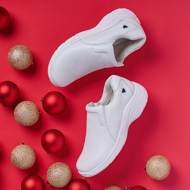 White Dove slip on shoes laid flat on a bright red background with Christmas bulbs decorating around the shoe. 